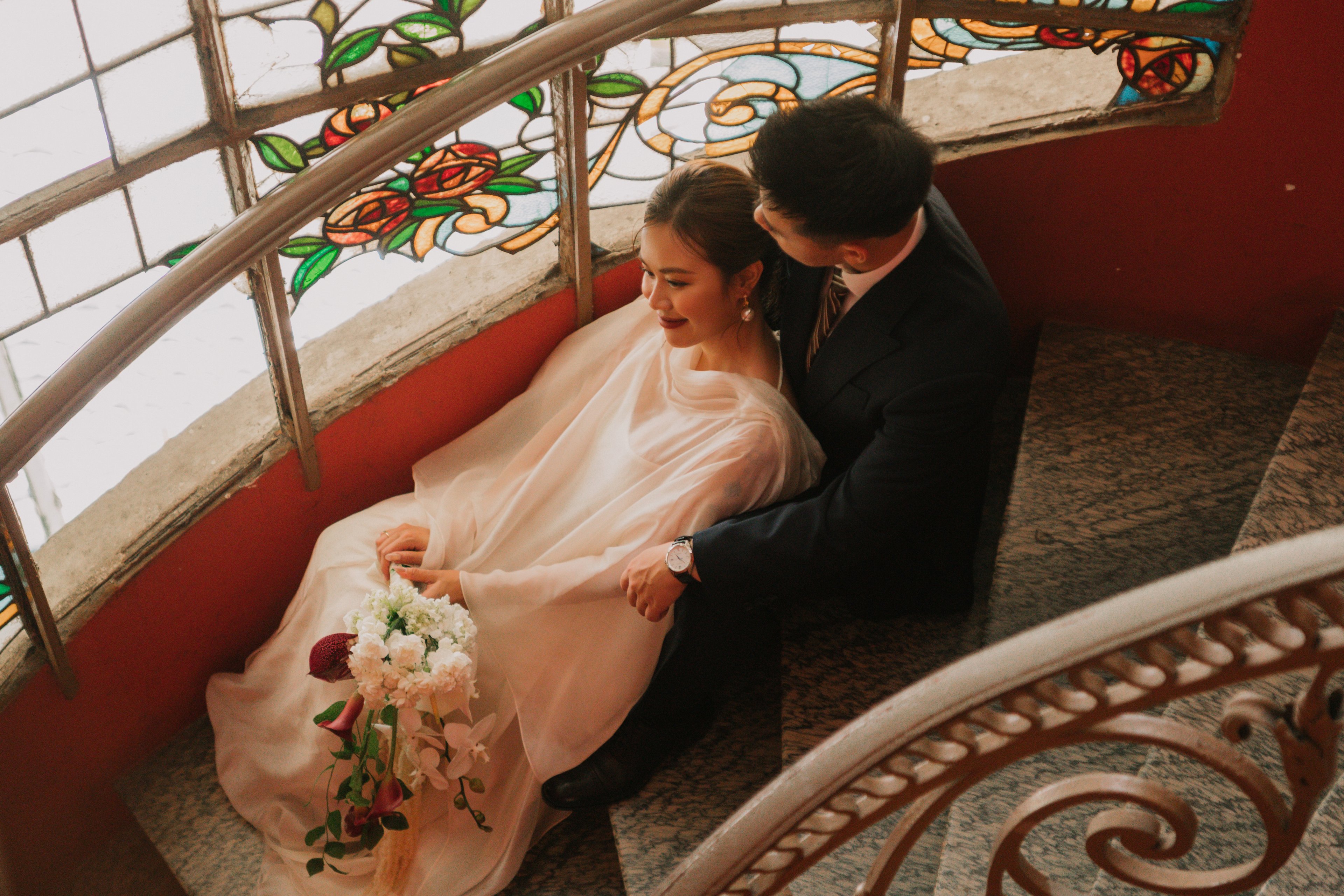 Ngọc Biết and Đức Vũ seated by a spiral staircase, stained glass behind them